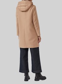 Manteau en laine beige avec une capuche matelassée, coupe droite, design superposé, porté avec un pantalon large en denim noir et des bottes Chelsea noires.