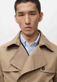 Beige trenchcoat met een brede kraag, met een dubbelzijdig ontwerp en een knoopaccent. Gedragen over een lichtblauw shirt.