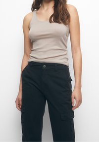 Žena v béžovém topu bez rukávů a černých kapsáčích, stojící proti jednobarevnému bílému pozadí.