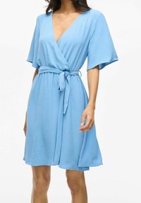 Woman wearing a knee-length light blue wrap dress with short wide sleeves and a tied waist belt, standing against a plain background.