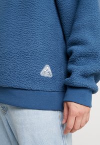 Blauer Fleece-Pullover mit strukturierter Oberfläche, gerippten Bündchen und einem dreieckigen Logo-Patch nahe dem Saum. Kombiniert mit hellblauen Jeans.
