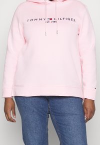 Vaaleanpunainen huppari, joka on tehty puuvillasekoitteesta. Siinä on kengurun tasku, säädettävä huppu ja logo, jossa lukee "TOMMY HILFIGER" ja "EST. 1985".