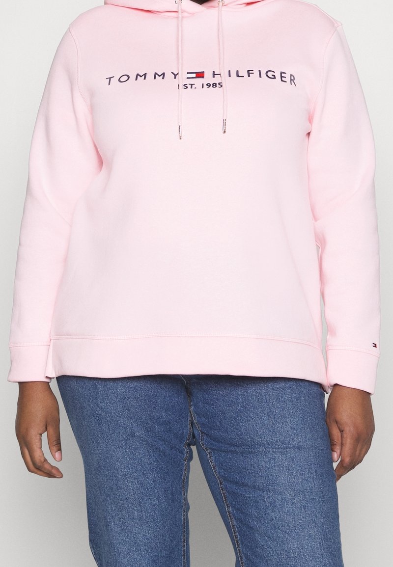 Vaaleanpunainen huppari, joka on tehty puuvillasekoitteesta. Siinä on kengurun tasku, säädettävä huppu ja logo, jossa lukee "TOMMY HILFIGER" ja "EST. 1985".