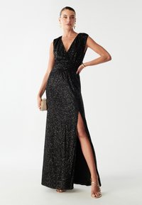 Sort palietkjole med dyb V-udskæring, korte ærmer og samlet talje. Den har en slidser op til låret og en svævende silhuet. Guld accessories fuldender looket.