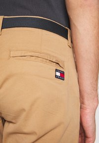 Beżowe spodnie z czarnym elastycznym pasem, z tylną kieszenią i naszywką logo "Tommy Jeans" w kolorze niebieskim, czerwonym i białym.