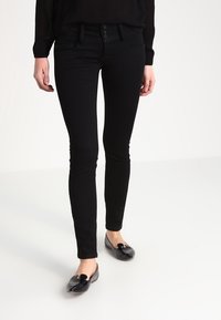 Persona vestida con jeans ajustados negros y bailarinas de charol negras, de pie sobre un suelo blanco contra un fondo blanco liso.