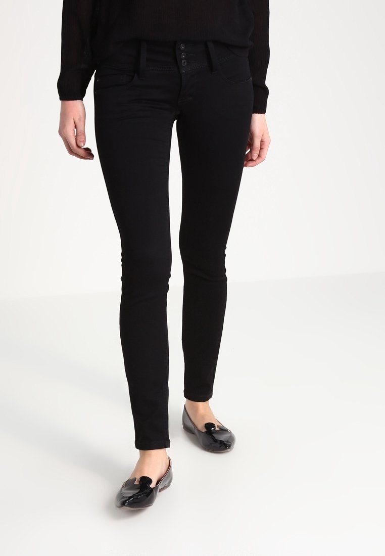 Persona vestida con jeans ajustados negros y bailarinas de charol negras, de pie sobre un suelo blanco contra un fondo blanco liso.