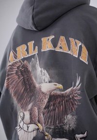 Felpa con cappuccio grigio scuro caratterizzata da una grande grafica di un'aquila in volo con montagne sullo sfondo e il testo "CARL KANI" stampato in giallo.