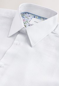 Chemise blanche texturée avec col pointu et doublure à motif floral à l'intérieur du col, partiellement boutonnée sur le devant.