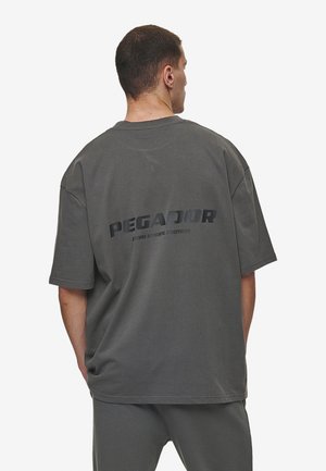 Grå, overdimensioneret t-shirt lavet af bomuld med korte ærmer. Har et dristigt print med "PEGADOR" i mørke bogstaver på ryggen. Glat tekstur.