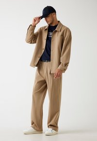 Camicia beige con bottoni, t-shirt blu navy, pantaloni beige sartoriali, sneaker bianche e un cappellino nero con logo, caratterizzati da una vestibilità rilassata e una texture liscia.
