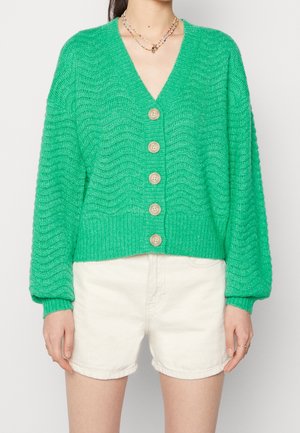 Kobieta ubrana w zielony, teksturowany kardigan z guzikami i długimi rękawami oraz białe szorty z wysokim stanem, uzupełnione koralikowymi naszyjnikami.