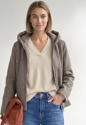 Femme aux cheveux blonds courts portant un pull beige à col en V, une veste grise zippée et un jean bleu, tenant un sac couleur rouille dans une main.