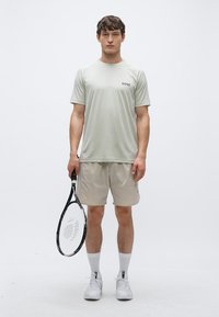 Ανοιχτό γκρι αθλητική μπλούζα με λογότυπο «BOSS», σε συνδυασμό με μπεζ σορτς. Το μοντέλο κρατάει μια μαύρη ρακέτα του τένις, φοράει λευκές κάλτσες και παπούτσια.