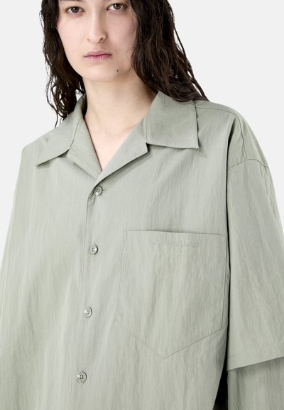 Persona con cabello largo y oscuro que lleva una camisa gris claro suelta abotonada, con un bolsillo en el pecho bordado con "CAMPERLAB".