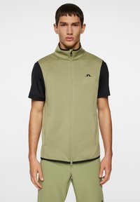 Ärmlös olivgrön väst med hög krage, framtill dragkedja och sidofickor, i kombination med svart kortärmad skjorta. Minimal varumärkesidentifikation synlig.