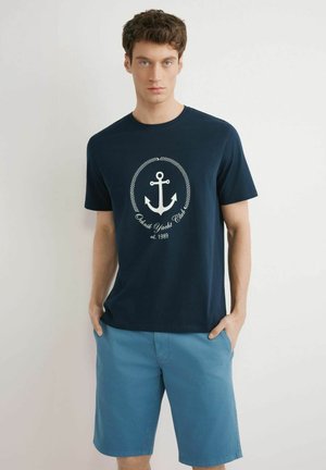 Młody mężczyzna w granatowej koszulce z nadrukiem kotwicy i jasnoniebieskich krótkich spodenkach, stoi z rękami w kieszeniach na jednolitym tle.