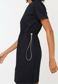 Kobieta w czarnej sukience z krótkimi rękawami, z regulowanym sznurkiem w talii, który ściąga materiał z boku.