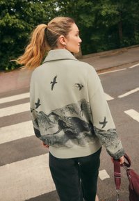 Jachetă oversized verde deschis cu guler, având imprimeuri cu păsări negre și munți pe spate. Asortată cu pantaloni închiși la culoare și o geantă bordeaux.