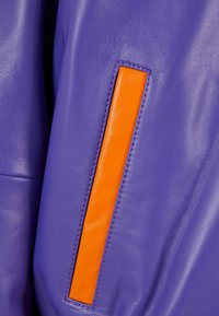 Fialová kožená látka má na boku diagonální oranžový kožený akcent. Textura je hladká s viditelnými detaily stehu.