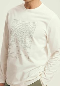 Biała koszulka z długim rękawem wykonana z bawełny, na przodzie z nadrukiem mapy topograficznej w odcieniach szarości. Materiał ekologiczny.