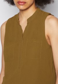 Top sem mangas de cor verde oliva com gola de bico e dois bolsos no peito. Tecido suave com uma textura lisa e detalhes de costura sutis.