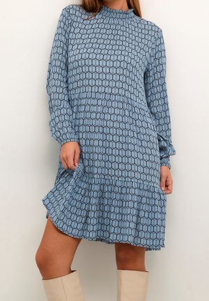 Femme portant une robe à manches longues bleue à motifs, cintrée à la taille avec un ourlet à volants, associée à des bottes beige montant jusqu'aux genoux.