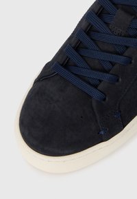 Prim-plan al vârfului unei tenăs de suede albastru închis, cu șireturi asortate și o talpă din cauciuc texturat, de culoare albă crem, pe un fundal deschis.