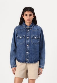 Cămașă din denim într-o nuanță medie de albastru, cu închidere prin nasturi, guler larg, două buzunare pe piept și cusături în contrast. Mâneci lungi, croi lejer.