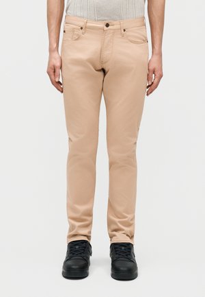 Άνδρας που φοράει μπεζ παντελόνι στενής γραμμής και μαύρα αθλητικά παπούτσια, στέκεται μπροστά σε απλό φωτεινό φόντο.