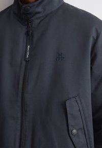 Jūrinė striukė, pagaminta iš lygaus audinio, su aukštu apykakle, užtrauktuku ir išsiuvinėtu logotipu. Šone yra spaudimo mygtukų detalė.