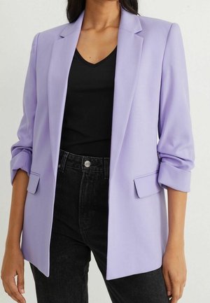 Kvinde iført en lys lilla blazer med opkørte ærmer, en sort V-hals top og sorte højtaljede jeans mod en neutral baggrund.
