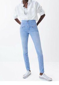 Jean en denim bleu clair taille haute avec poches avant, jambes étroites et fermeture par bouton, associé à une chemise blanche et des baskets blanches.