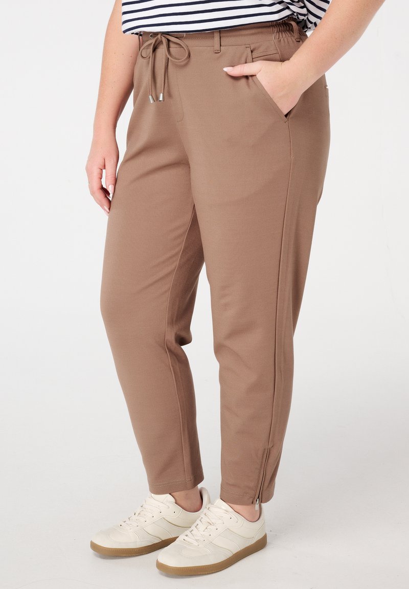 Pantalons marron cintrés avec taille à cordon, poches latérales et zips aux chevilles. Fabriqués en tissu doux, associés à des baskets blanches.