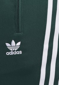 Temno zeleni športni hlače s belim tridelnim dizajnom, ki vključujejo žep z zadrgo in vezeno belo adidas logo.