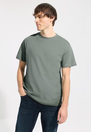 Grünes Baumwoll-T-Shirt mit rundem Ausschnitt und kurzen Ärmeln, präsentiert an einer männlichen Figur mit dunklen Jeans. Glatte Textur, lässige Passform.