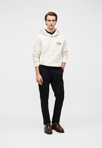 Εκρού φούτερ με κουκούλα, εμπρόσθια τσέπη και λογότυπο, συνδυασμένο με μαύρο παντελόνι και καφέ παπούτσια. Το μοντέλο στέκεται μπροστά σε απλό φόντο.