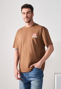 Brun bomulds t-shirt med korte ærmer og rund hals. Forsiden har et pink solgrafik og ordet "SOLAR" i hvid. Båret med denimjeans.