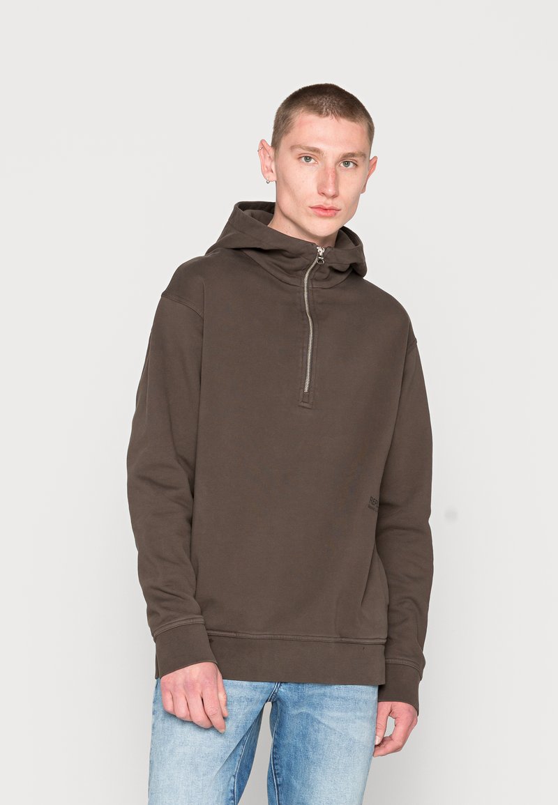 Brauner Kapuzenpullover mit halber Reißverschlussfront, lockerer Passform, gerippten Bündchen und Saum. Getragen mit hellblauen Jeans und minimalistischer Markenkennzeichnung.