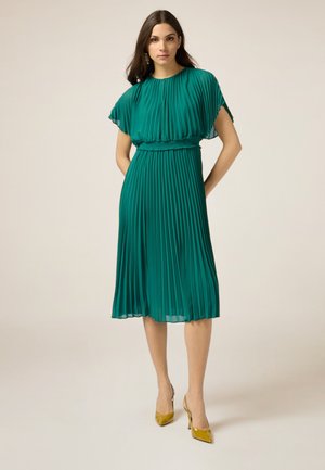 Turquoise geplisseerde jurk met korte cape mouwen, een cinched taille en een vloeiende rok. Gecombineerd met gele hoge hakken.