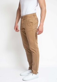 Hombre vestido con chinos beige ajustados, zapatillas blancas y una camiseta blanca metida por dentro, de pie frente a un fondo blanco liso.