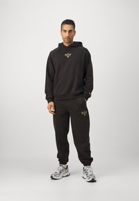 Black hoodie and matching sweatpants made of cotton blend, featuring a gold emblem on the chest and thigh. Rubber-soled athletic shoes.