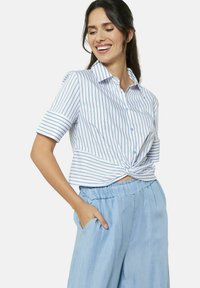 Camicia button-up accorciata bianca con righe verticali blu; presenta maniche corte arrotolate e un dettaglio anteriore annodato. Abbinata a pantaloni blu chiaro.