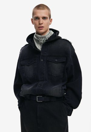 Schwarze Denim-Übergröße-Jacke mit zwei Fronttaschen, fransigen Schultern, abgenutztem Material, kombiniert mit einem grauen gemusterten Schal und einem schwarzen Gürtel.