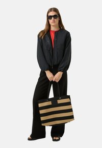 Czarna kurtka bomberka, czerwona koszula, czarne spodnie i duże okulary przeciwsłoneczne. Trzyma beżowo-czarną torbę typu tote z czarnymi uchwytami i okuciami.