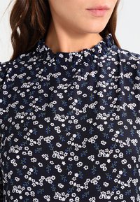 Marinblus med ett blommigt mönster av vita och blå blommor, med en volangkrage och lätt tyg.