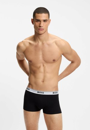Homme torse nu aux cheveux courts portant un caleçon boxer noir BOSS, debout les mains derrière le dos sur un fond blanc uni.