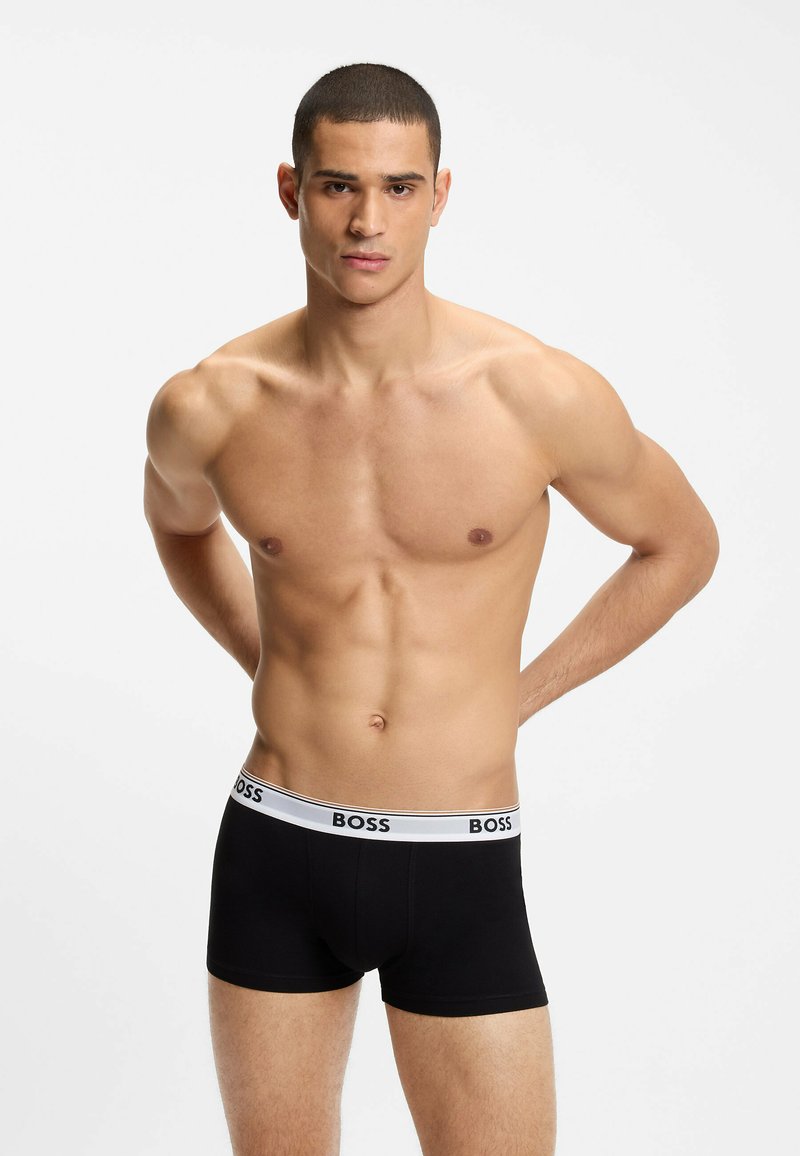 Homme torse nu aux cheveux courts portant un caleçon boxer noir BOSS, debout les mains derrière le dos sur un fond blanc uni.