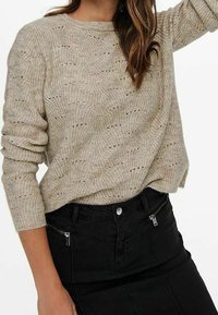 Beige gestrickter Pullover mit strukturiertem Design, rundem Ausschnitt und langen Ärmeln, kombiniert mit einem schwarzen, taillierten Rock mit einer Reißverschlusstasche.
