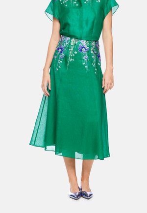 Grünes Midikleid mit lila und weißer Blumenstickerei um die Taille, transparenter Stoff, getragen mit metallisch lila spitzen Schuhen.